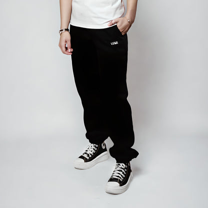 Women's LCC Sweatpants Black shown on model against light background, featuring relaxed fit, elastic cuffs, small LUMI logo, and paired with black and white sneakers. Side view showcasing comfortable fit and classic jogger style.