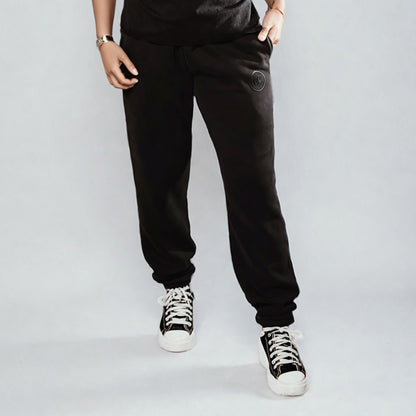 Women's LUX Sweatpants Black Wash shown on model against light background, featuring relaxed fit with cuffed ankles, elastic waistband, and raised rubber logo detail. Paired with black and white sneakers, showcasing the comfortable cotton-blend fabric in a versatile black wash finish.