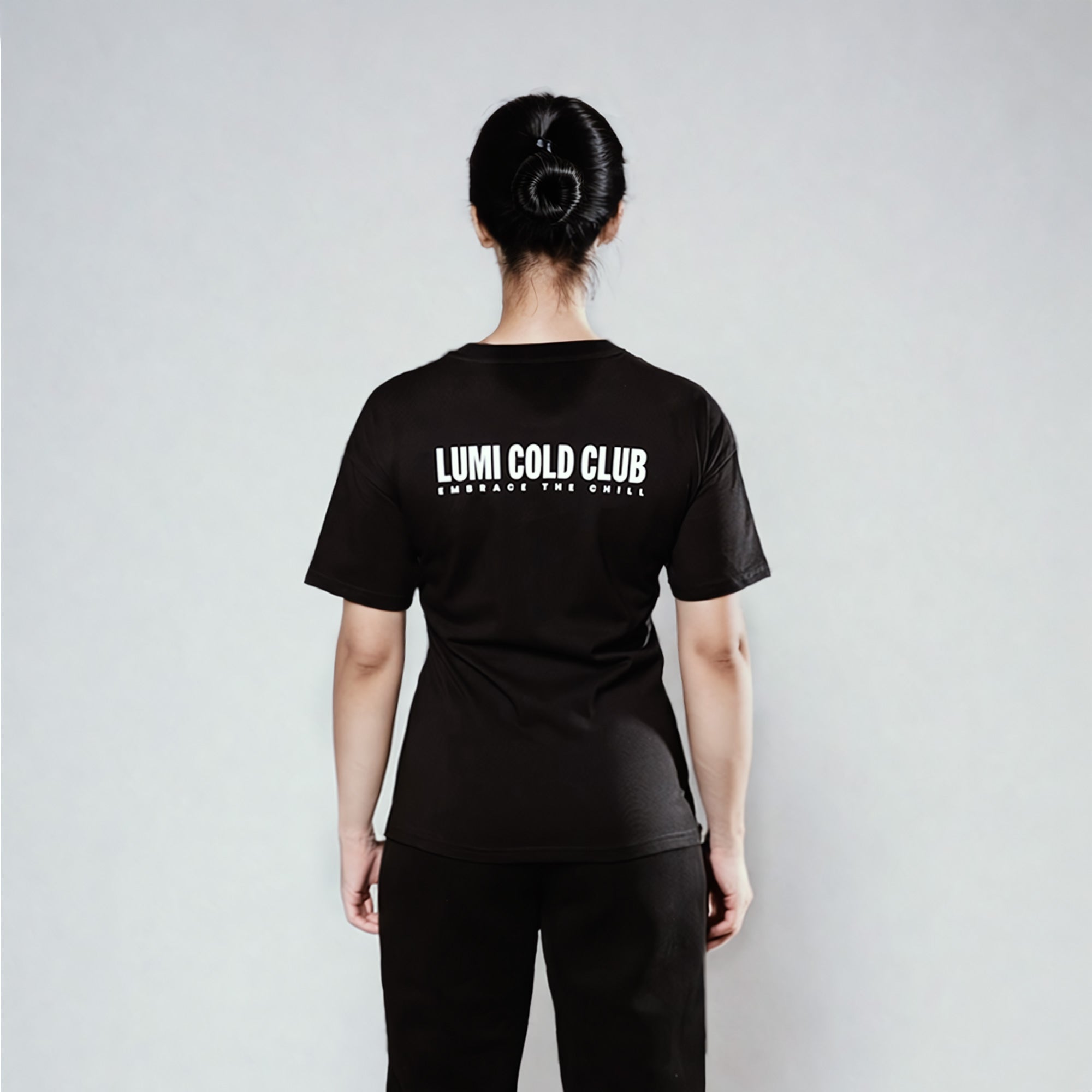 Back view of Women's LCC T-Shirt Black showing LUMI Cold Club text graphic in white, styled with black pants against a light gray background. Regular fit heavyweight cotton t-shirt with classic crew neck design.