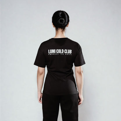 Back view of Women's LCC T-Shirt Black showing LUMI Cold Club text graphic in white, styled with black pants against a light gray background. Regular fit heavyweight cotton t-shirt with classic crew neck design.