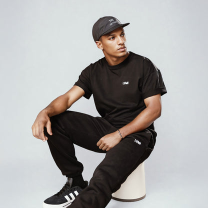 LUMI Cold Bundle showcased on a model wearing the black oversized t-shirt and matching joggers, paired with a black cap and athletic shoes, sitting casually against a light gray background, demonstrating the comfortable, athleisure style of the collection.