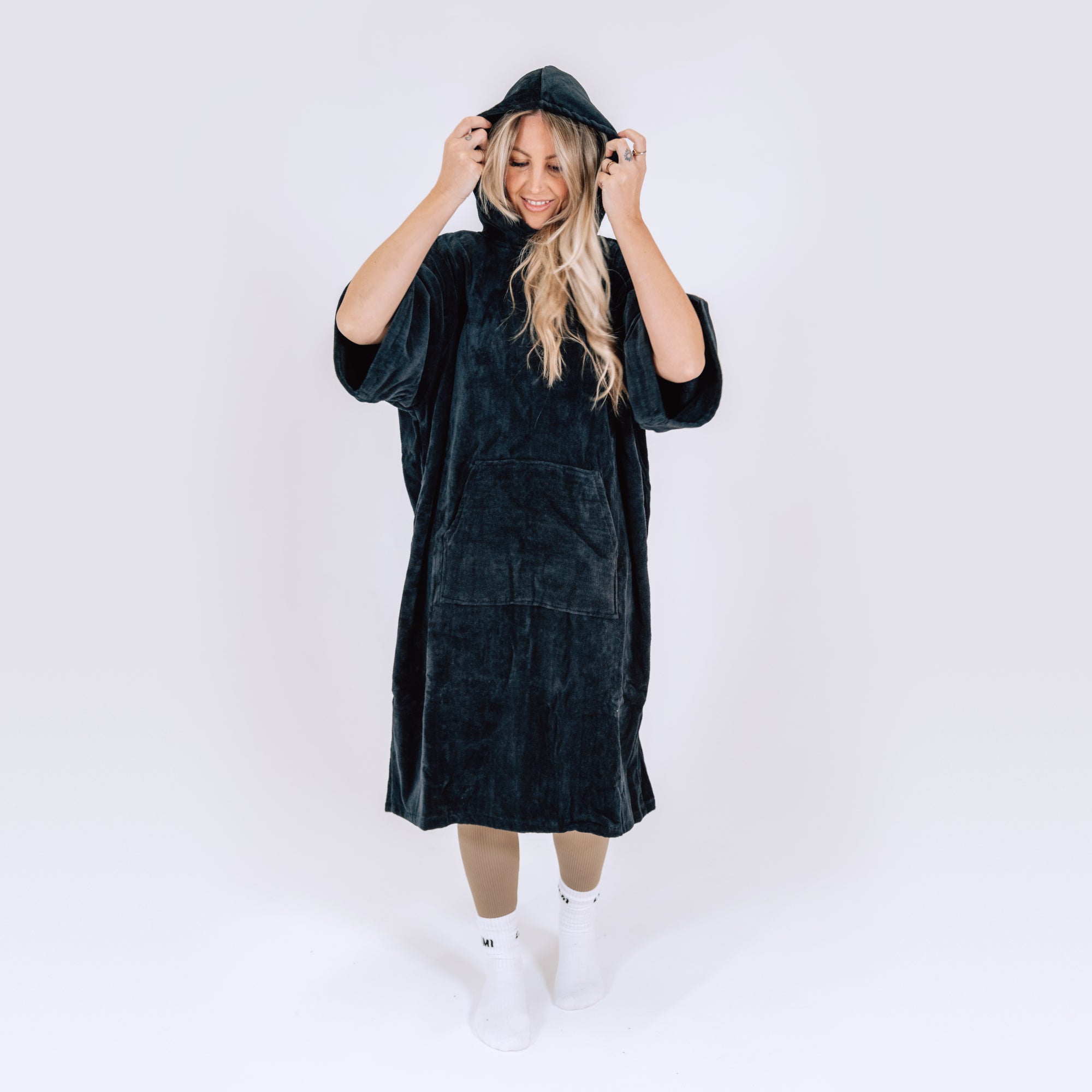 LUMI Towel Poncho shown in black plush cotton fabric, featuring oversized hooded design with front pocket, modeled against white background. Knee-length robe demonstrates roomy fit and cozy comfort perfect for post-swim or ice bath use.