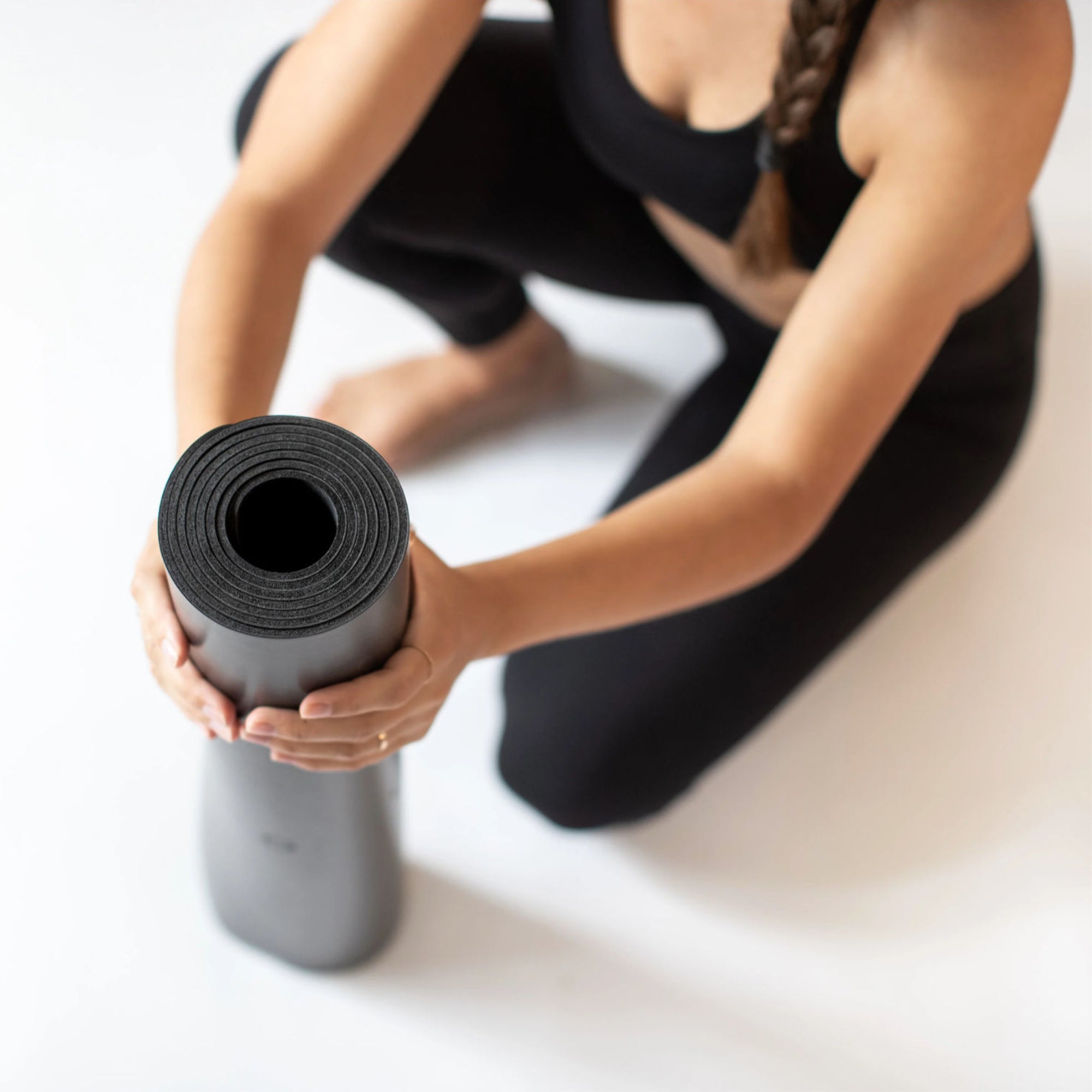 A person in black athletic wear rolling up an Eco Wolf Yoga Mat in Matcha color, sitting on white background. Top-down view showing hands carefully handling the premium non-slip exercise mat.