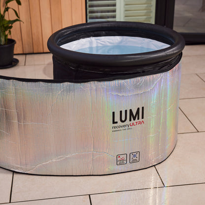 LUMI Ultra Cover, an iridescent thermal cover for ice baths, featuring a holographic silver finish and black trim, displayed on beige tile flooring next to a potted plant, showing its innovative insulating wrap-around design with filled water basin